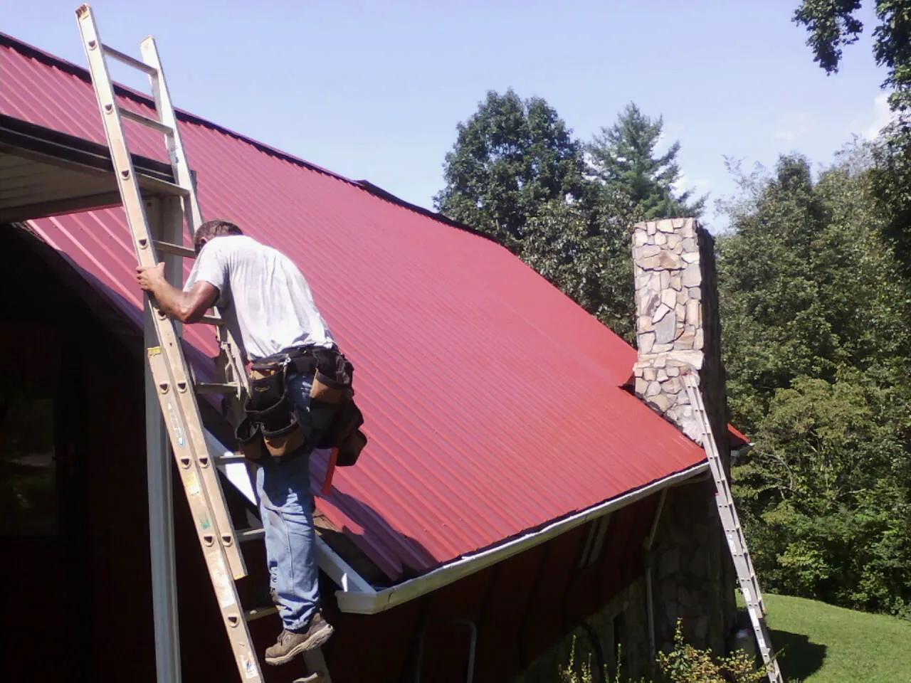 Roofer on a ladder beside a red metal roof and stone chimney — Soffit & Fascia Repair by Roofing Brothers in Crescent